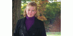 Frau mit blondem Haar im Freien vor einem Baum, trägt violetten Rollkragen und schwarze Jacke.