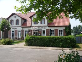 Gasthaus mit roten Ziegeldächern und gepflegten Hecken, umgeben von Bäumen bei sonnigem Wetter.