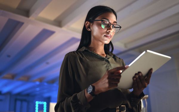 Eine Mitarbeiterin einer Firma steht in einem Großraumbüro und blickt auf ein Tablet in ihrer Hand. Ihre Augen schützt sie mit einer Bürobrille.