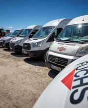 Mehrere weiße Transporter mit "FUCHS"-Logo parken nebeneinander auf einem Schotterplatz.