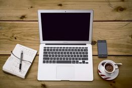 Laptop, notitieboek, pen, telefoon en koffiekopje op houten tafel.