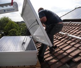 Hombre instalando paneles solares en un tejado inclinado de tejas marrones.