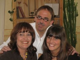 Eine Gruppe von drei Personen lächelt in einer wohnlichen Umgebung mit Pflanzen und Bücherregal.