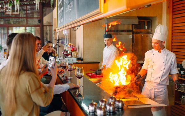 Zwei Köche stehen in einer offenen Küche. Einer der beiden Köche flambiert etwas mit einer hohen Flamme. Die Gäste können von ihren Plätzen aus zuschauen und halten ihre Handys in der Hand.