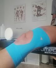 Rodilla vendada con cinta azul en una consulta médica. Pósteres anatómicos de fondo.