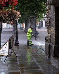 Persona pulisce il marciapiede con l'idropulitrice; cartello di pulizia sul lato.