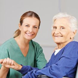 Caregiver in green scrubs smiling with an elderly woman in a wheelchair, both holding hands.