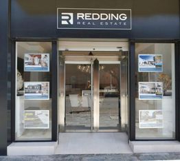 Fachada de oficina de bienes raíces con escaparate de propiedades y puertas de cristal.