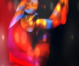 Person tanzt in buntem Licht, trägt ein rotes Outfit und hat die Arme über den Kopf ausgestreckt.