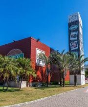 Prédio vermelho com palmeiras na frente e torre alta exibindo logotipos de lojas, sob céu azul.