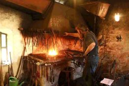 Schmied arbeitet in Werkstatt, umgeben von Werkzeugen, mit Feuer in der Esse.