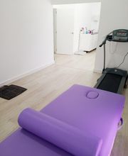 Sala blanca con camilla morada y caminadora; puerta abierta al fondo con vista al pasillo.