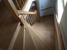 Holztreppe mit Geländer windet sich abwärts, Licht fällt durch ein Fenster.