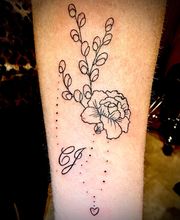 Een tatoeage met bloemen, takjes, initialen "C J" en een klein hartje op een onderarm.