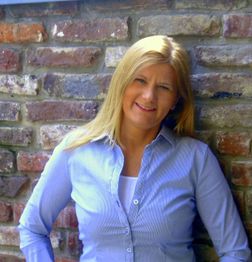 Eine lächelnde Frau in einem blauen Hemd lehnt an einer Backsteinwand.