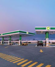 Gasolinera al atardecer con dos bombas y vehículos estacionados, paisaje montañoso al fondo.