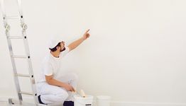 Painter kneeling by a ladder, pointing at a blank wall, with paint supplies nearby.
