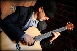 Ein Mann mit Mütze spielt Gitarre vor einer Backsteinwand.