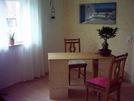 Zimmer mit Holztisch, zwei Stühlen und Pflanze. Licht fällt durch ein Fenster mit weißen Vorhängen.