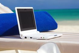 Laptop auf Liegestuhl am Strand mit blauem Handtuch, Meer im Hintergrund.