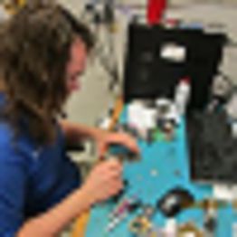 Person repairing electronics at a cluttered workbench with various tools and components.