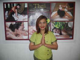 Person in grüner Kleidung vor einem Poster mit Bildern von Thai-Massagen, lächelnd mit gefalteten Händen.