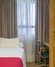 Quarto simples com cama de solteiro, escrivaninha e estante de madeira ao lado da janela.