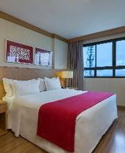 Quarto de hotel com cama de casal, roupa de cama branca, manta vermelha e janela com cortinas.