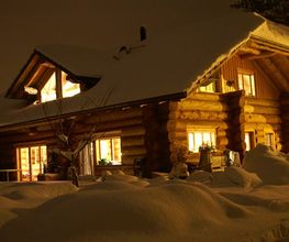 Verschneites Blockhaus bei Nacht, warme Beleuchtung im Inneren, dicker Schnee bedeckt das Dach.
