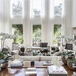 Sala elegante com janelas altas, sofás claros, plantas e livros sobre a mesa de centro.