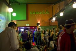 Grupo de pessoas em um bar movimentado, com placa "casaclub" ao fundo. Ambiente descontraído.