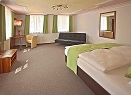 Helles Hotelzimmer mit Doppelbett, Sofa, Sessel, TV, Spiegel, grünen Vorhängen und Teppichboden.