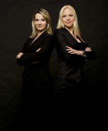 Zwei Frauen in schwarzen Anzügen stehen Rücken an Rücken und verschränken die Arme vor schwarzem Hintergrund.