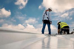 Two workers on a rooftop, wearing safety gear, checking equipment under a blue sky with clouds.