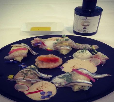 Piatto gourmet con pesce, fiori ed erbe su piatto nero. Bottiglia di olio a lato.
