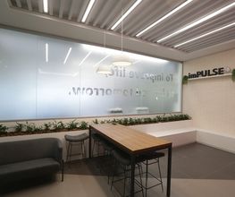 Sala moderna com mesa alta, bancos e sofá. Placas de vidro com letreiro e plantas decorativas.