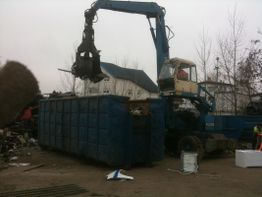 Ein blauer Kran lädt Schrott in einen großen Container auf einem Industriegelände.