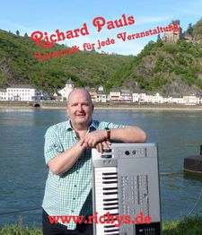 Ein Mann steht mit einem Keyboard am Fluss, im Hintergrund eine Burg und Bäume.