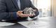 A person in a suit holds a model house above insurance documents and keys on a table.