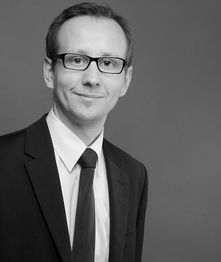 Ein Mann in Anzug und Krawatte, trägt Brille, vor grauem Hintergrund, lächelt leicht. Schwarzweißfoto.