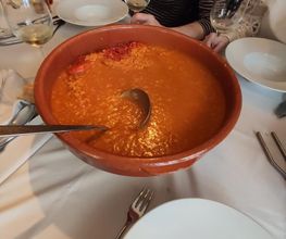 Cuenco de arroz caldoso en mesa con platos, cubiertos, y copas vacías. Personas al fondo.