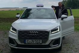 Ein Mann steht neben einem weißen Audi mit Fahrschul-Schild, auf einem Feldweg.