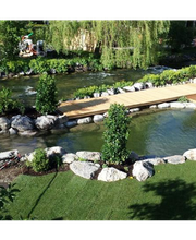 Holzbrücke über Teich in grüner Gartenlandschaft mit Bäumen und Felsen.