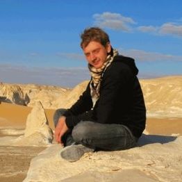 Eine Person sitzt lächelnd auf einem Felsen in einer Wüstenlandschaft unter blauem Himmel.