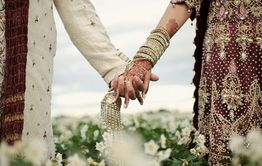 Couple in ornate attire holding hands in a field of flowers, showing henna and intricate designs.