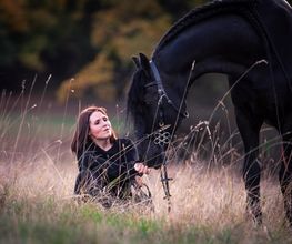 Frau sitzt in einem Feld neben einem großen, schwarzen Pferd. Sie hält dessen Zügel, während sie es anschaut.