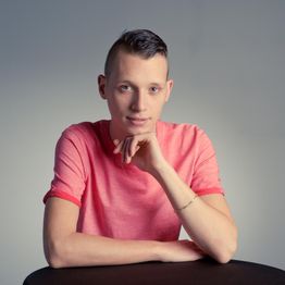 Jeune homme en t-shirt rose assis, regardant la caméra avec le menton appuyé sur sa main.