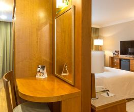 Quarto de hotel com cama, TV, mesa com espelho e cadeiras. Cortinas compridas ao fundo.