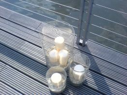 Kerzen in Glasgefäßen auf Holzplanken neben einem Wasserbereich.