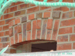 Ziegelmauer mit dekorativem Bogenfenster, von grüner Abdeckung teilweise verdeckt.